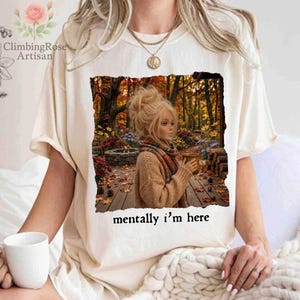 May include: A cream-colored short-sleeve t-shirt featuring a graphic print of a person with blonde hair in a bun, wearing a tan sweater and scarf, holding a mug in an autumn forest setting. The text below the graphic reads, mentally i'm here.