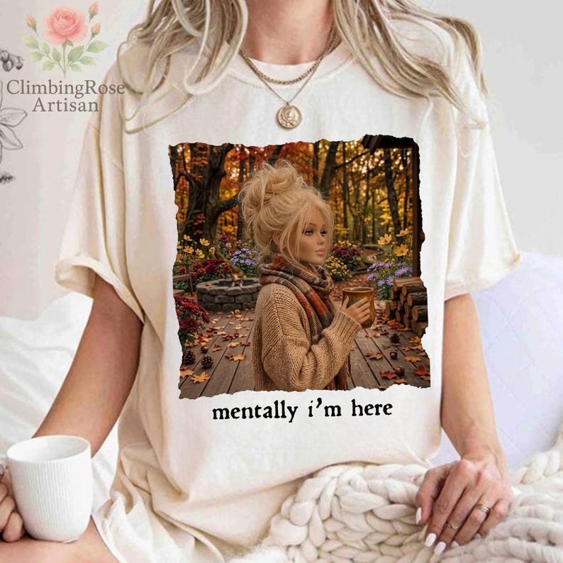 May include: A cream-colored short-sleeve t-shirt featuring a graphic print of a person with blonde hair in a bun, wearing a tan sweater and scarf, holding a mug in an autumn forest setting. The text below the graphic reads, mentally i'm here.