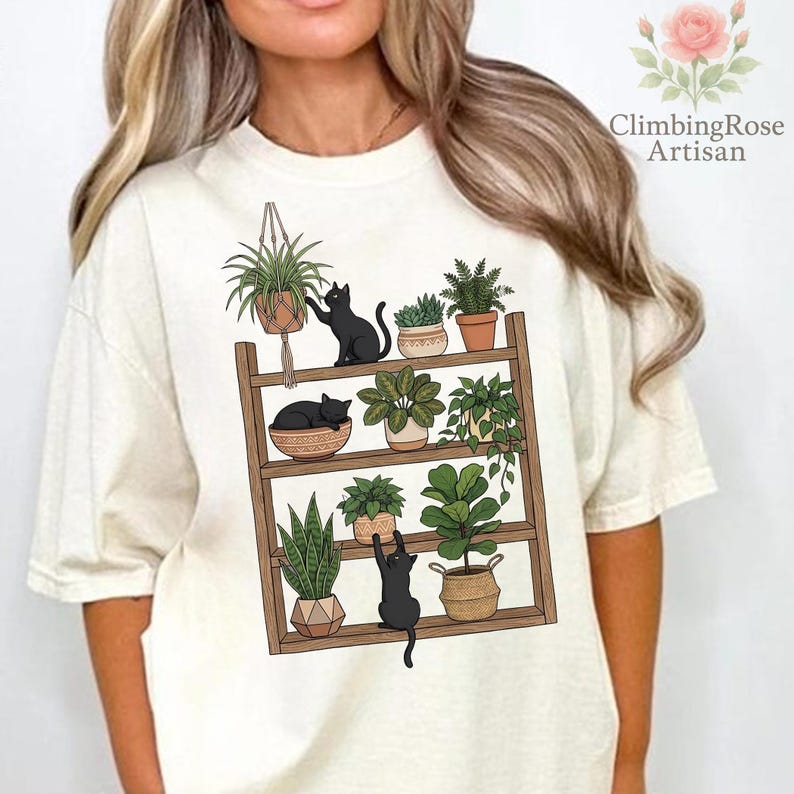 May include: Cream-colored t-shirt featuring a graphic of a wooden plant shelf with various potted plants and three black cats. The shelf has three levels, with the cats interacting with the plants. The text "ClimbingRose Artisan" is on the upper right.