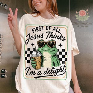 May include: A cream-colored t-shirt featuring a graphic of a green frog wearing leopard-print heart-shaped sunglasses and holding an iced coffee. The text reads: First of all, Jesus Thinks I'm a delight. The design includes a black and white checkerboard pattern.