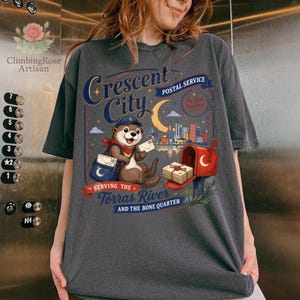 May include: Dark gray t-shirt featuring a whimsical illustration of an otter in a postal worker uniform. The design includes the text "Crescent City Postal Service" and "Serving the Torras River and the Bone Quarter."