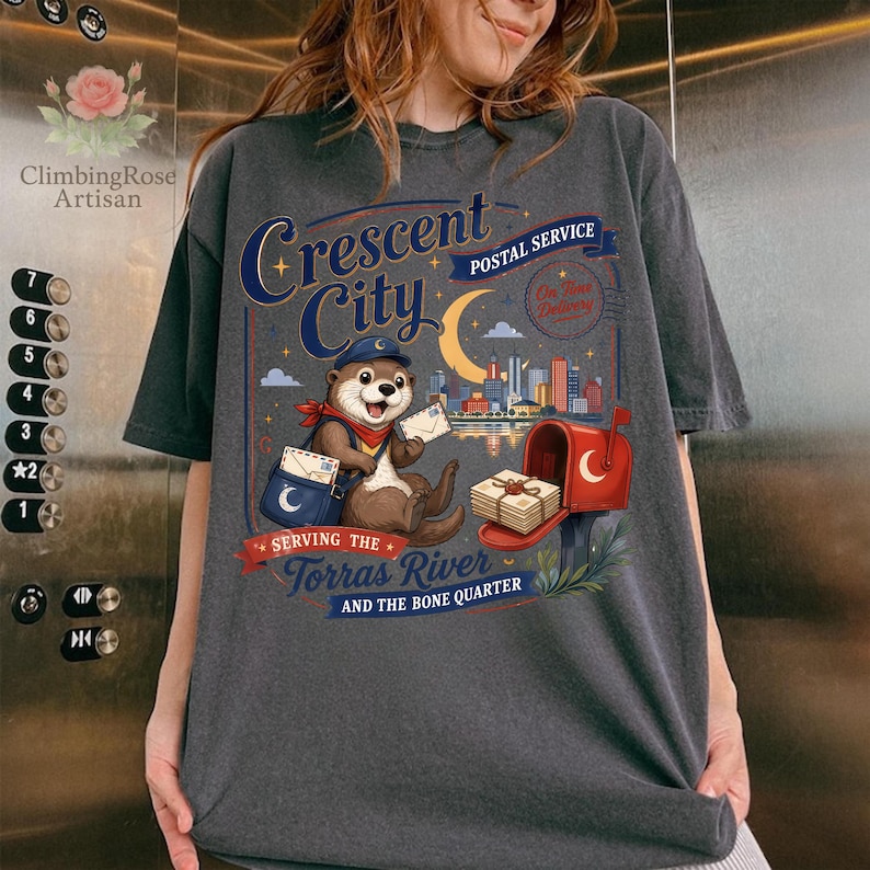 May include: Dark gray t-shirt featuring a whimsical illustration of an otter in a postal worker uniform. The design includes the text "Crescent City Postal Service" and "Serving the Torras River and the Bone Quarter."