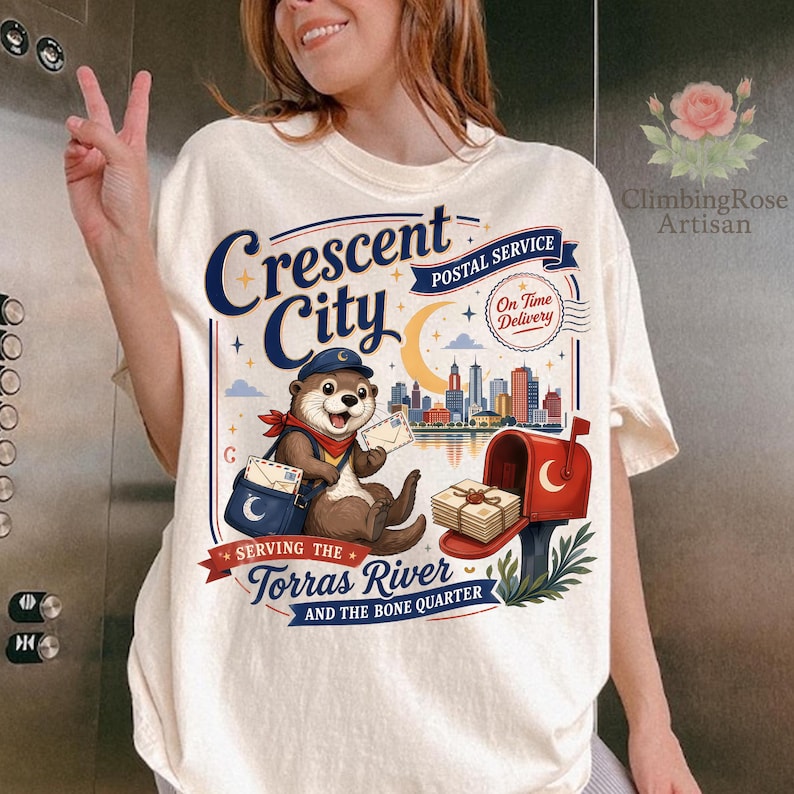 May include: Cream-colored t-shirt featuring a graphic of an otter in a postal worker uniform, holding letters. The design includes the text "Crescent City Postal Service" and "Serving the Torras River and the Bone Quarter."