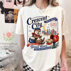 May include: Cream-colored t-shirt featuring a whimsical illustration of an otter in a postal worker uniform. The design includes the text "Crescent City Postal Service" and a cityscape. The shirt also has the text "Serving the Torras River and the Bone Quarter."