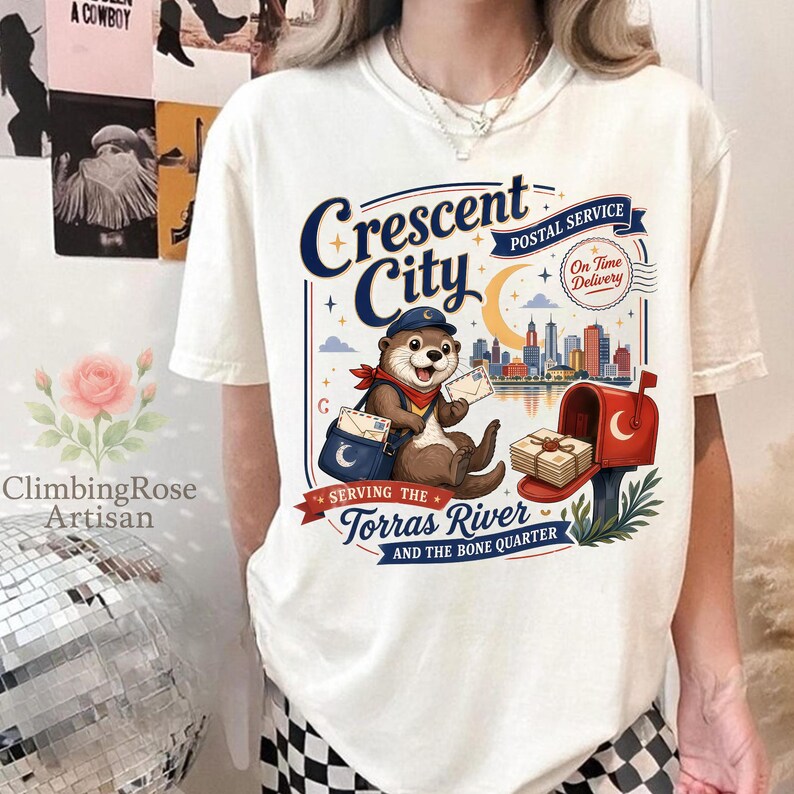 May include: Cream-colored t-shirt featuring a whimsical illustration of an otter in a postal worker uniform. The design includes the text "Crescent City Postal Service" and a cityscape. The shirt also has the text "Serving the Torras River and the Bone Quarter."