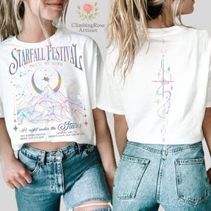 May include: White t-shirt with a colorful graphic design. The front features the text "STARFALL FESTIVAL" above a mountain scene with a crescent moon. The back has a vertical design with stars and crescent moons.