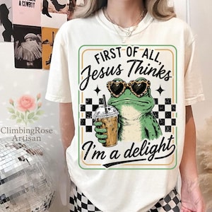 May include: A cream-colored t-shirt featuring a graphic of a green frog wearing leopard-print heart-shaped sunglasses and holding an iced coffee. The text reads: First of all, Jesus Thinks I'm a delight, framed by a black and white checkerboard pattern.
