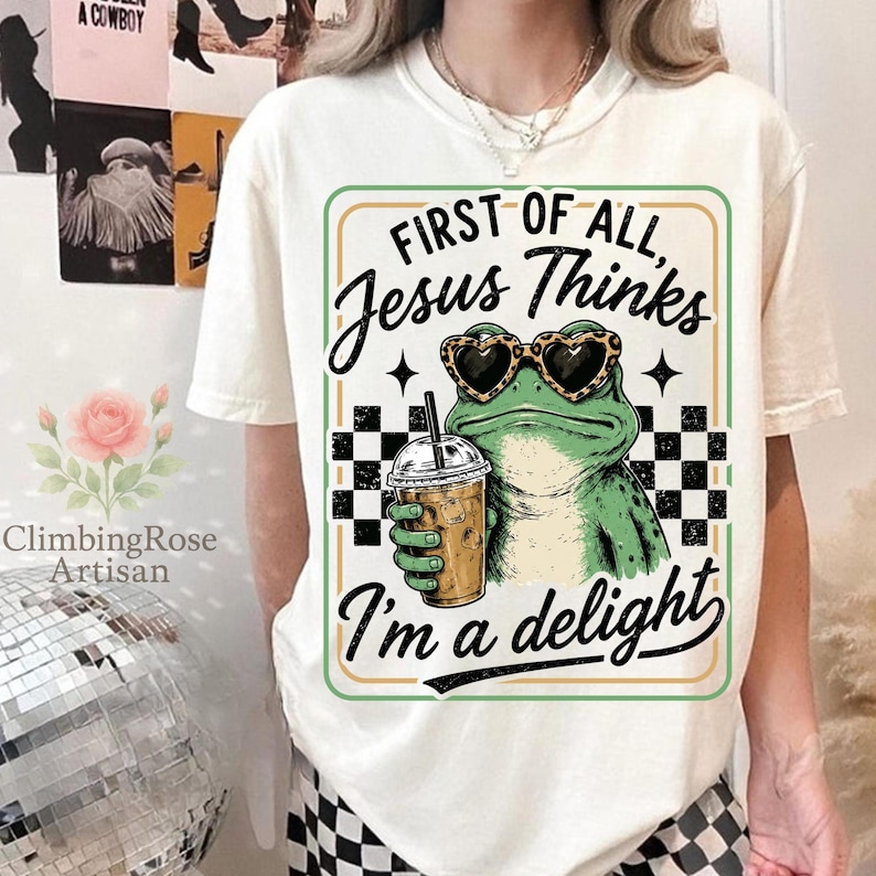 May include: A cream-colored t-shirt featuring a graphic of a green frog wearing leopard-print heart-shaped sunglasses and holding an iced coffee. The text reads: First of all, Jesus Thinks I'm a delight, framed by a black and white checkerboard pattern.