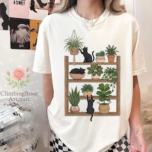 May include: Cream-colored t-shirt featuring a graphic of a wooden shelf with potted plants and black cats. The shelf holds various plants in decorative pots. One cat reaches for a hanging plant, while another rests in a woven basket.