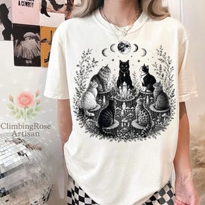 May include: An off-white short-sleeve t-shirt featuring a black ink graphic of six cats sitting on tree stumps in a circle around a central crystal cluster, with a moon phase cycle above and surrounding botanical elements like vines and mushrooms.