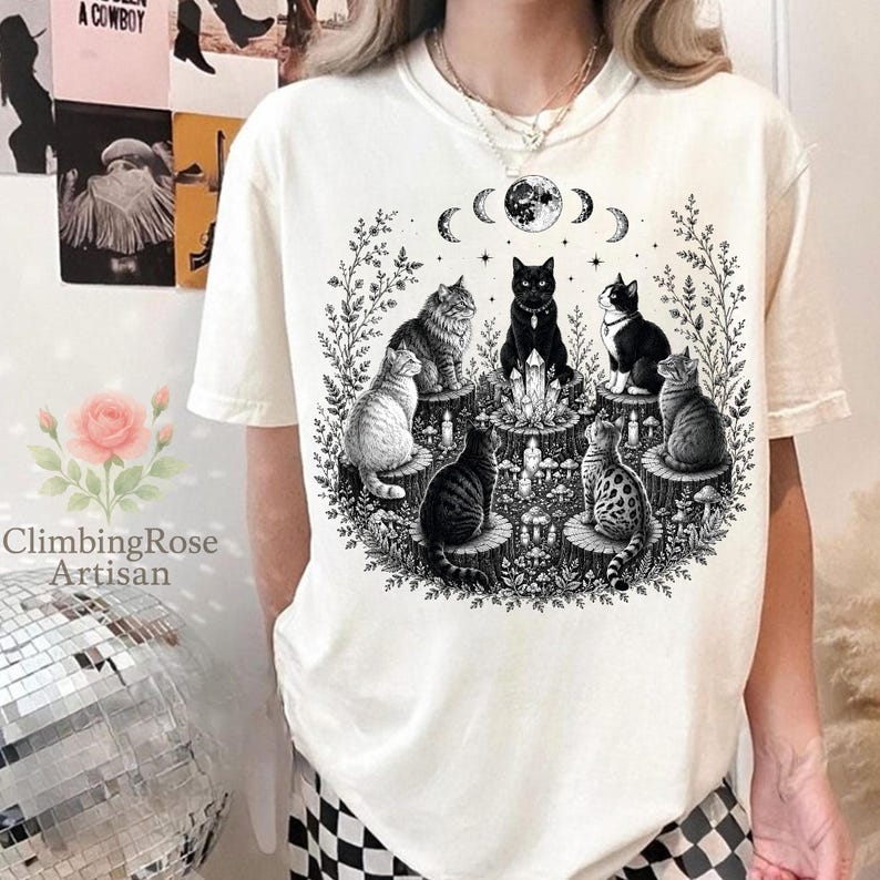 May include: An off-white short-sleeve t-shirt featuring a black ink graphic of six cats sitting on tree stumps in a circle around a central crystal cluster, with a moon phase cycle above and surrounding botanical elements like vines and mushrooms.