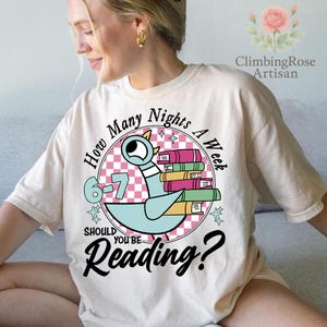 May include: Cream-colored t-shirt featuring a cartoon pigeon holding books, with the text "How Many Nights A Week Should You Be Reading?" in a circular design. The design includes a pink and white checkered background and the numbers 6-7.