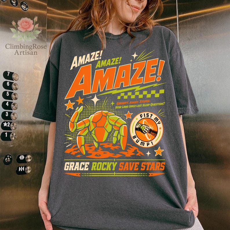 K&ouml;nnte beinhalten: Dunkelgraues T-Shirt mit einem Retro-Grafikdesign. Das Design zeigt das Wort "AMAZE!" in Orange und Wei&szlig;, einen Cartoon-Krebs und den Satz "FIST MY BUMP!". Das Shirt enth&auml;lt auch den Text "GRACE ROCKY SAVE STARS".