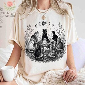 May include: A cream-colored short-sleeve t-shirt featuring a black ink-style graphic of six cats sitting on tree stumps in a circle around a central crystal formation, with a moon phase cycle above and surrounding botanical foliage elements.