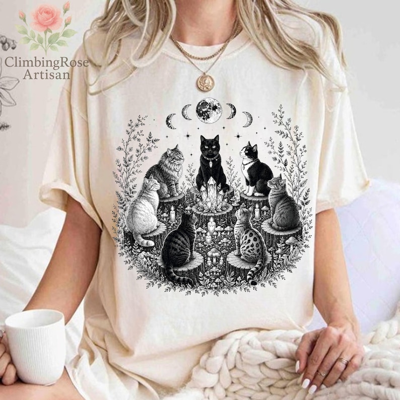 May include: A cream-colored short-sleeve t-shirt featuring a black ink-style graphic of six cats sitting on tree stumps in a circle around a central crystal formation, with a moon phase cycle above and surrounding botanical foliage elements.