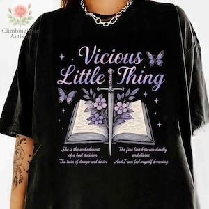Może przedstawiać: Czarny T-shirt z fioletowym i białym nadrukiem graficznym przedstawiającym książkę z wystającą z niej szpadą. Tekst "Vicious Little Thing" znajduje się nad książką. Tekst pod książką brzmi: "She is the embodiment of a bad decision. The twin of danger and desire. The fine line between deadly and divine. And I can feel myself drowning."