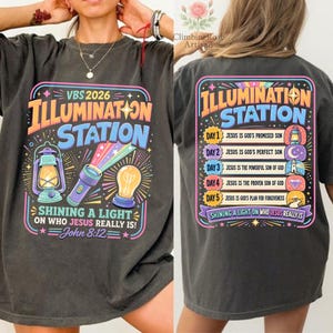 Pu&ograve; includere: T-shirt grigio scuro con un design grafico colorato. La parte anteriore presenta il testo "VBS 2026 ILLUMINATION STATION" con illustrazioni di una lanterna, una torcia e una lampadina. Il retro ha lo stesso testo e un elenco di temi giornalieri.
