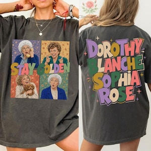 Puede incluir: Camiseta gris oscuro con un gráfico colorido de las Golden Girls. La parte delantera muestra las palabras "Stay Golden" con imágenes de los personajes. La parte trasera de la camiseta tiene los nombres Dorothy, Blanche, Sophia y Rose.