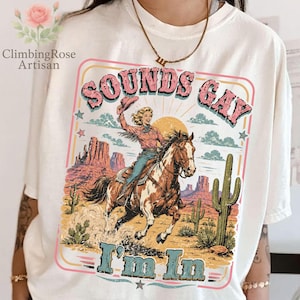 Puede incluir: Camiseta color crema con un diseño retro de una vaquera montando a caballo en un paisaje desértico. El texto "SOUNDS GAY I'M IN" se muestra en una fuente vintage. El diseño incluye un atardecer, cactus y estrellas.