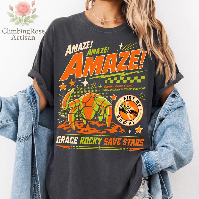 K&ouml;nnte beinhalten: Dunkelgraues T-Shirt mit einem Retro-Grafikdesign. Das Design zeigt das Wort "AMAZE!" in Orange und Gelb, ein krabbenartiges Wesen und den Text "FIST MY BUMP!". Zus&auml;tzlicher Text lautet "GRUMPY, ANGRY, STUPID. HOW LONG SINCE LAST SLEEP QUESTION?" und "GRACE ROCKY SAVE STARS".