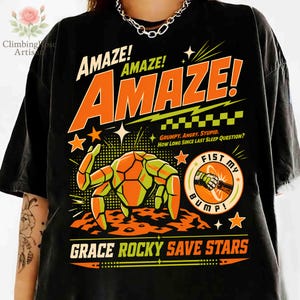 K&ouml;nnte beinhalten: Schwarzes T-Shirt mit einem Retro-Grafikdesign. Das Design zeigt das Wort "AMAZE!" in gro&szlig;en orangefarbenen Buchstaben, eine geometrische Krabbe und den Satz "FIST MY BUMP!". Zus&auml;tzlicher Text enth&auml;lt "GRUMPY. ANGRY. STUPID. HOW LONG SINCE LAST SLEEP QUESTION?" und "GRACE ROCKY SAVE STARS."