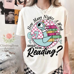 May include: Cream-colored t-shirt with a graphic featuring a blue pigeon holding books, surrounded by text that reads "How Many Nights A Week Should You Be Reading?" The design includes a pink and white checkered circle.