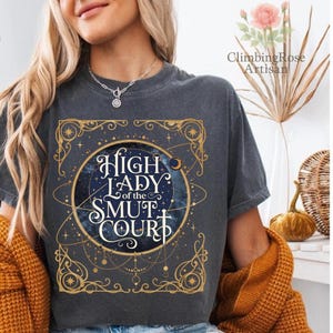 May include: Dark gray t-shirt with a celestial design featuring the text "High Lady of the Smut Court" in a decorative font. The design includes a starry night sky and gold accents. The shirt is worn by a person.