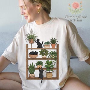 May include: Cream-colored t-shirt featuring a graphic of a wooden shelf with potted plants and black cats. The shelf holds various plants in pots and hanging baskets. The text "ClimbingRose Artisan" is printed on the upper right.