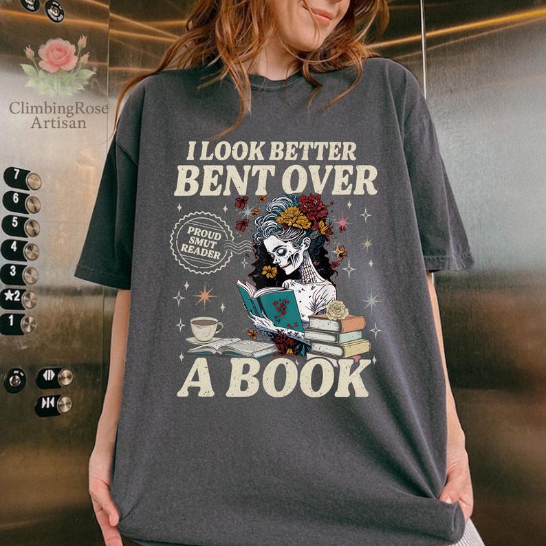 May include: Dark gray t-shirt with the text "I LOOK BETTER BENT OVER A BOOK." The graphic features a skeleton reading a book, with flowers in their hair. A coffee cup and books are also in the design. The shirt has a vintage, worn look.