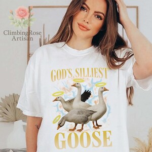 May include: White t-shirt with the text "GOD'S SILLIEST GOOSE" in gold lettering. The graphic features three geese with halos, set against a soft, colorful background. The shirt is a casual, comfortable style.