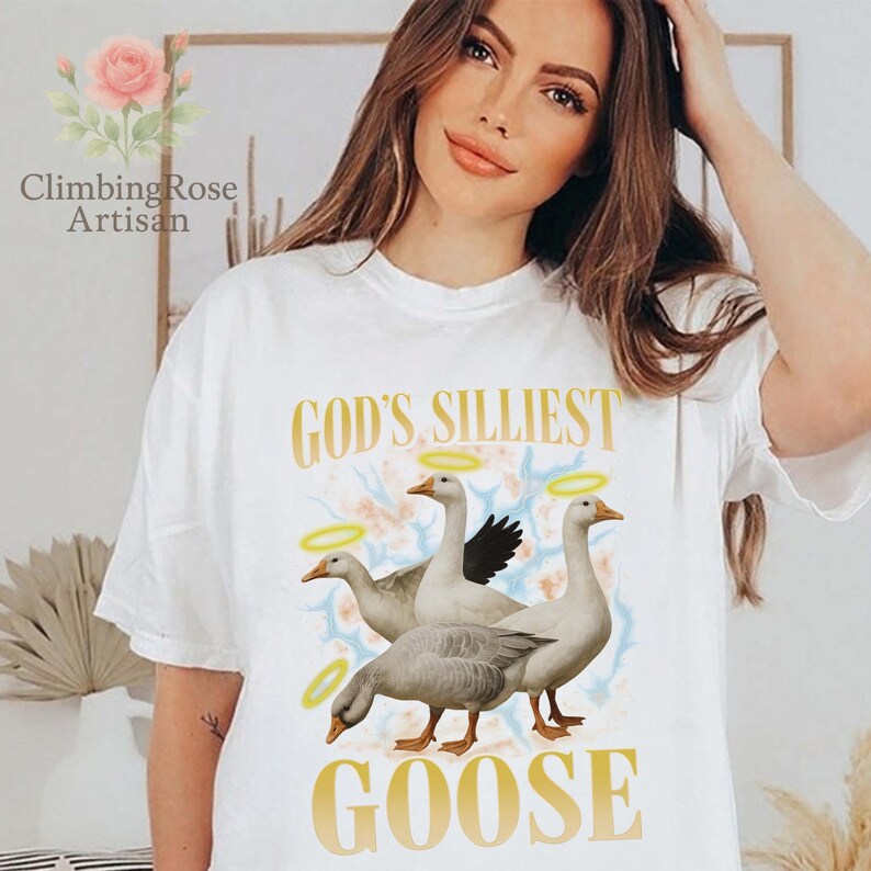 May include: White t-shirt with the text "GOD'S SILLIEST GOOSE" in gold lettering. The graphic features three geese with halos, set against a soft, colorful background. The shirt is a casual, comfortable style.
