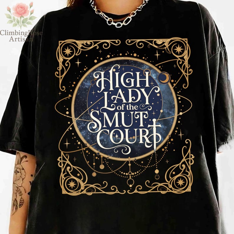 May include: Black t-shirt featuring a celestial design with the text "High Lady of the Smut Court" in a decorative font. The design includes stars, a crescent moon, and ornate gold borders. The shirt is a casual wear item.