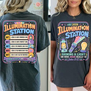 Pu&ograve; includere: T-shirt grigio scuro con un design grafico colorato. Il design presenta il testo "ILLUMINATION STATION" e "VBS 2026", insieme a illustrazioni di una lanterna, una torcia e una lampadina. La maglietta include anche il testo "Shining a Light on Who Jesus Really Is!" e "John 8:12".