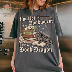 Pode incluir: T-shirt cinzenta escura com um gr&aacute;fico que diz "I'm Not A Bookworm, I'm A Book Dragon". O design apresenta uma pilha de livros, flores e um drag&atilde;o lendo um livro. O logotipo Climbing Rose Artisan est&aacute; no canto superior esquerdo.