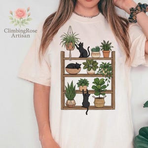 May include: Cream-colored t-shirt featuring a graphic of a wooden shelf filled with potted plants and black cats. The shelf holds various plants, including hanging plants, succulents, and leafy greens. The text "ClimbingRose Artisan" is on the left side.