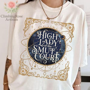 May include: Cream-colored t-shirt featuring a celestial design with the text "High Lady of the Smut Court" in a decorative font. The design includes a blue and gold color scheme with stars and ornate borders. The shirt is a casual wear item.