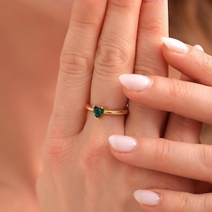 Może przedstawiać: Złoty pierścionek z ciemnozielonym kamieniem szlachetnym w kształcie serca. Pierścionek ma prosty design, z cienką obrączką i wyeksponowanym kamieniem szlachetnym. Pierścionek jest noszony na palcu.