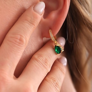 May include: Gold hoop earrings featuring a teardrop-shaped, emerald-green gemstone. The hoops are embellished with small, clear crystals. The image shows the earring in the ear of a person.