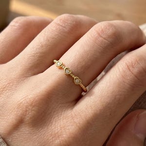 May include: A gold-colored ring featuring a series of heart-shaped designs. The ring is delicate and appears to be made of a thin band with small, heart-shaped embellishments. The ring is worn on a finger.