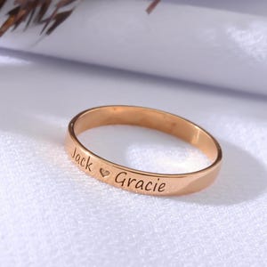 Può includere: Un anello in oro rosa con i nomi "Jack" e "Gracie" incisi, separati da un cuore. L'anello è posto su uno sfondo bianco con accenti floreali secchi.