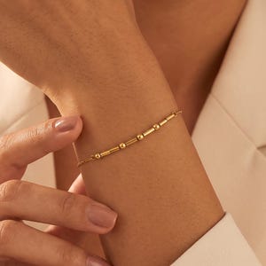 May include: A delicate gold-toned chain bracelet featuring a series of small, cylindrical beads and round spheres arranged in a linear pattern. The bracelet is secured with a thin, polished metal chain that wraps around the wrist.