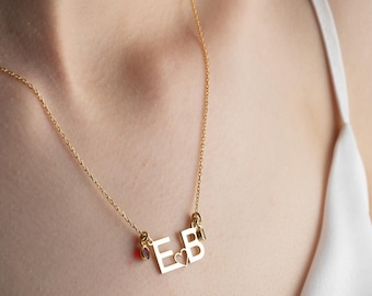 May include: A gold-colored necklace with the letters "E" and "B" is displayed. The letters are connected by a delicate chain. Small red gemstones and a small clear gemstone are attached to the letters. The necklace is worn on a person.