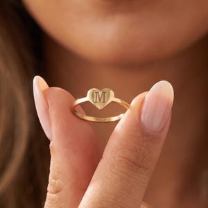 Könnte beinhalten: Ein goldener Ring mit einem herzförmigen Mittelstück, in dessen Mitte der Buchstabe M eingraviert ist. Das Ringband ist dünn und kreisförmig und besteht aus poliertem, goldfarbenem Metall. Das Design ist schlicht und betont das Herzmotiv.