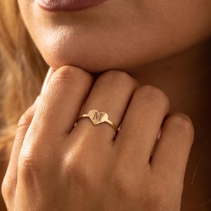 Könnte beinhalten: Ein goldener Ring mit einem herzförmigen Anhänger, in dessen Mitte der Buchstabe M eingraviert ist. Der Ring hat eine dünne, polierte Ringschiene und wird an einem Finger getragen. Das Design ist schlicht und metallisch mit einem klaren M.