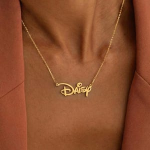 Op de afbeelding: Een goudkleurige ketting met de naam "Daisy" in een gestileerd lettertype. De hanger hangt aan een fijne ketting. De ketting wordt getoond tegen een bruine achtergrond.