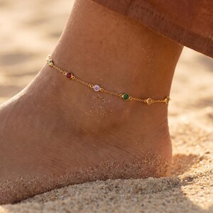 Op de afbeelding: Een enkelbandje van een goudkleurige ketting met kleine, ronde, in een zetting geplaatste edelstenen in diverse kleuren, waaronder rood, lichtpaars, groen en transparant. De fijne metalen ketting omsluit de enkel met gelijkmatig verdeelde stenen.