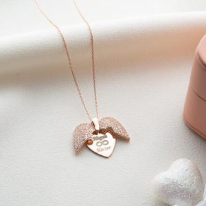 May include: Rose gold necklace with a heart-shaped pendant and wing accents. The pendant features the names "Megan" and "Kecter" with an infinity symbol. The necklace is displayed on a white fabric surface, next to a pink jewelry box.