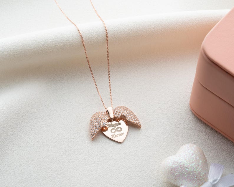 May include: Rose gold necklace with a heart-shaped pendant and wing accents. The pendant features the names "Megan" and "Kecter" with an infinity symbol. The necklace is displayed on a white fabric surface, next to a pink jewelry box.