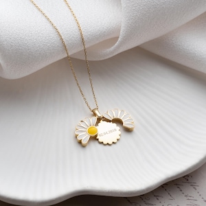 Puede incluir: Un collar con colgante de margarita de color dorado. El colgante presenta un dise&ntilde;o de margarita con p&eacute;talos de esmalte blanco y un centro amarillo. Tambi&eacute;n se adjunta un peque&ntilde;o dije redondo con texto grabado. El collar se muestra sobre una superficie blanca.