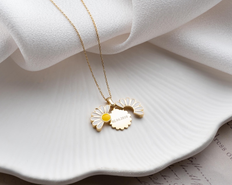 Puede incluir: Un collar con colgante de margarita de color dorado. El colgante presenta un dise&ntilde;o de margarita con p&eacute;talos de esmalte blanco y un centro amarillo. Tambi&eacute;n se adjunta un peque&ntilde;o dije redondo con texto grabado. El collar se muestra sobre una superficie blanca.